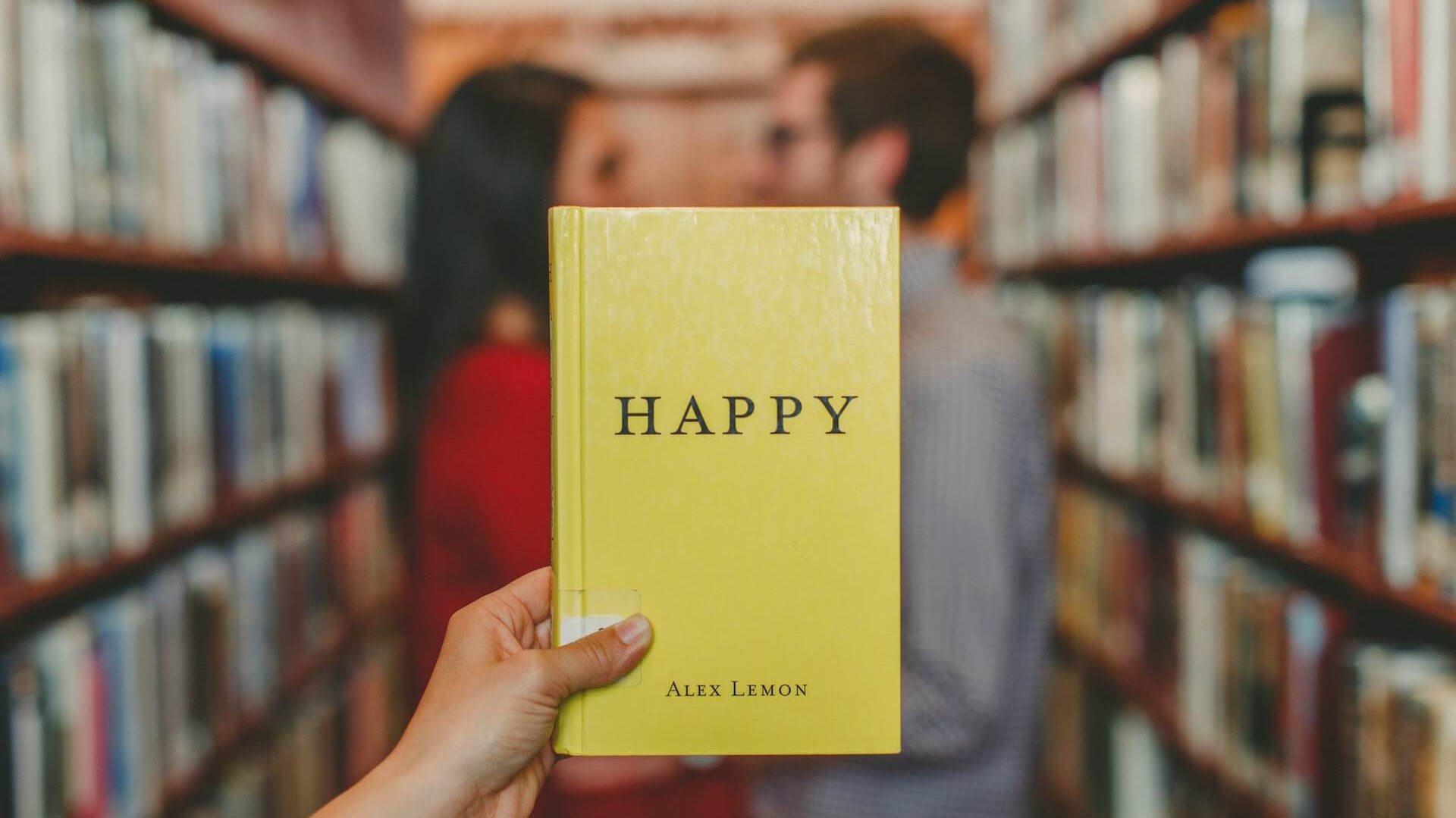 Scena di biblioteca con una coppia sullo sfondo e una persona che tiene in mano un libro giallo intitolato 'Happy'